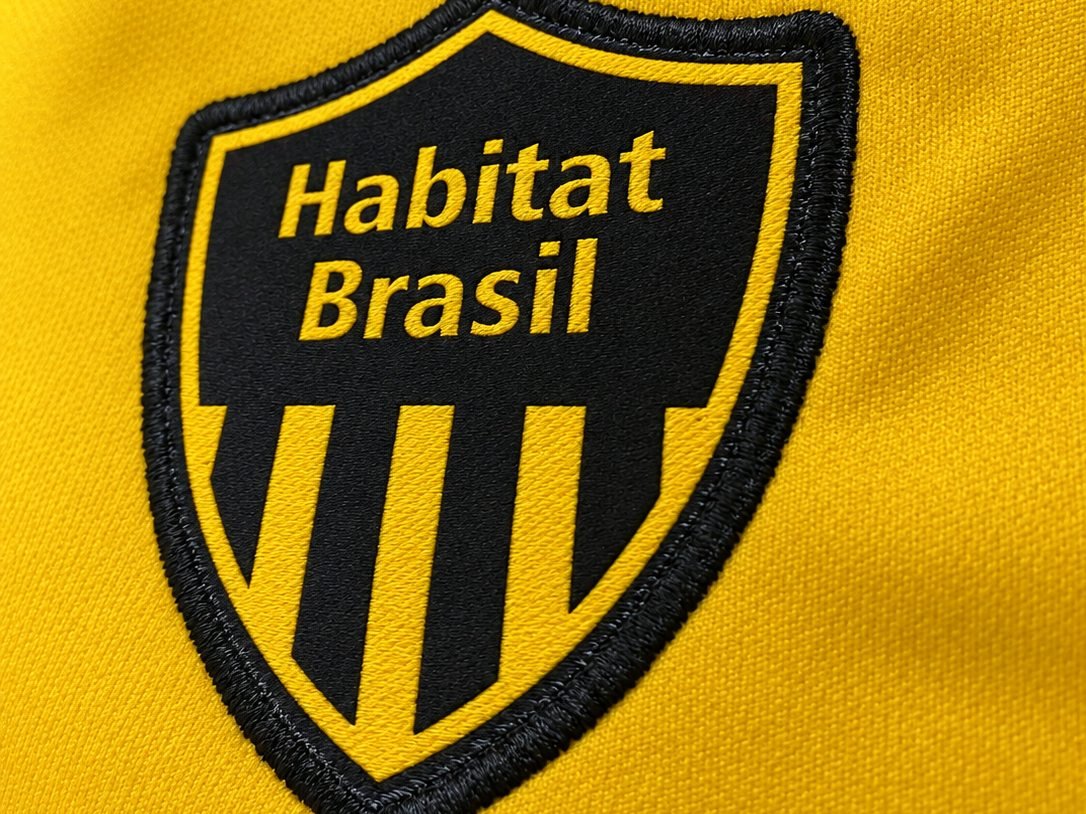 Close do bordado em camisa amarela: escudo Habitat Brasil com borda bordada em preto, mostrando o relevo dos pontos