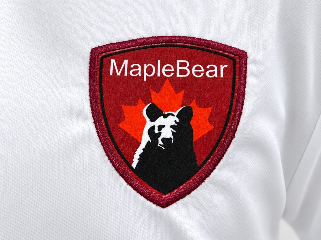 Close do bordado em camisa branca: escudo Maple Bear com borda bordada em vermelho, mostrando o relevo dos pontos