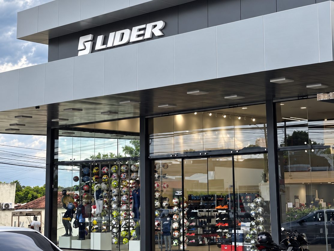 Entrada da loja da Lider Sport: letreiro Lider sobre a vitrine de vidro, com bolas e uniformes expostos