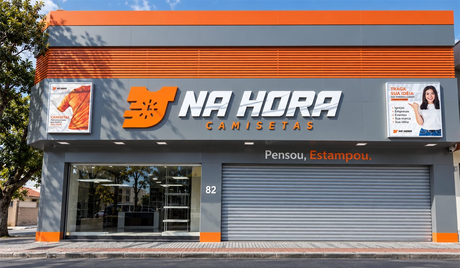 Fachada da loja da Na Hora Camisetas: prédio cinza com faixa laranja, o logo Na Hora Camisetas e a frase Pensou, Estampou. acima das vitrines